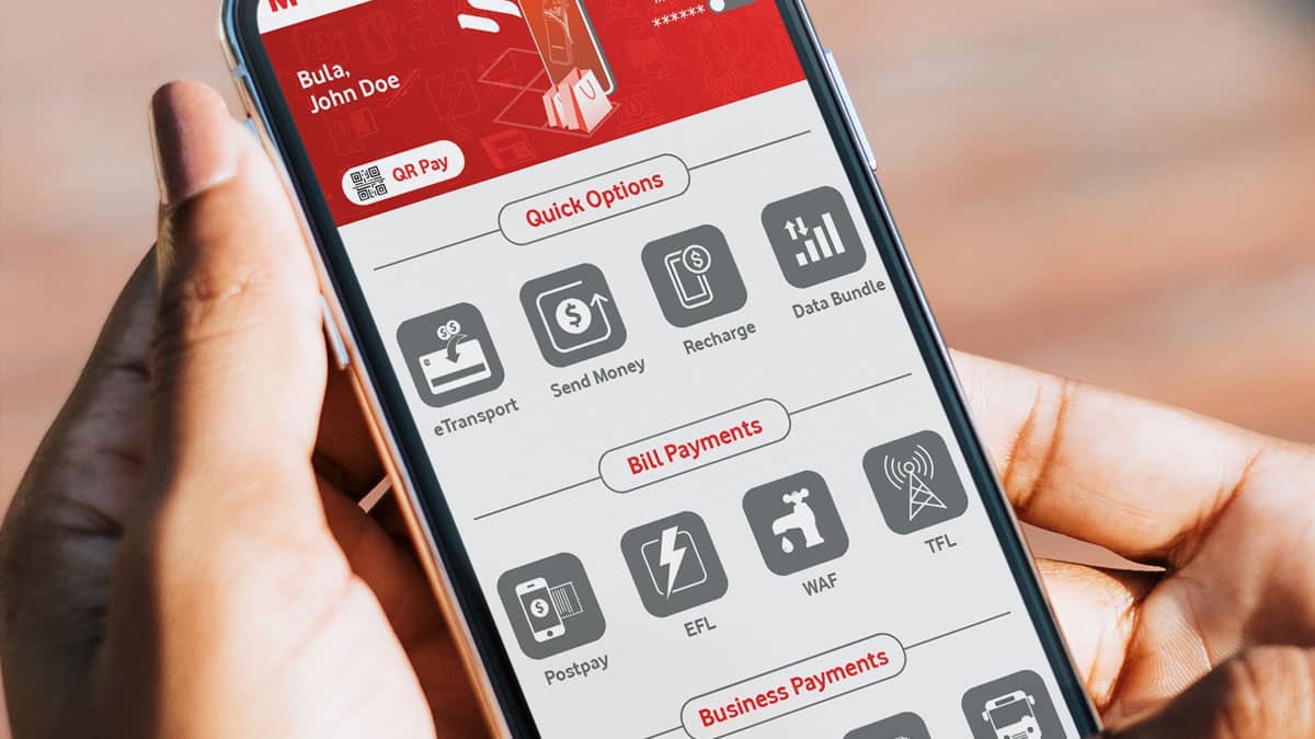 A person's hand holding a smartphone displaying the M-PAiSA mobile wallet app interface. The screen shows a red header with 'Bula, John Doe' and various service options organized in sections including Quick Options (e-Transport, Send Money, Recharge, Data Bundle), Bill Payments (Postpay, EFL, WAF, TFL), and Business Payments. The app interface features a QR Pay option and multiple gray icons representing different financial services.