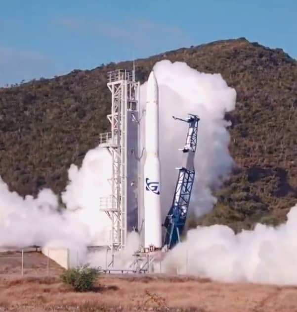 A white rocket stands upright on a launch pad, surrounded by support structures and releasing large clouds of white vapor or smoke. The rocket has a blue logo on its side. The scene is set outdoors at the base of a green, bushy hill under a clear sky. The ground around the pad is dry with sparse grass and shrubs.