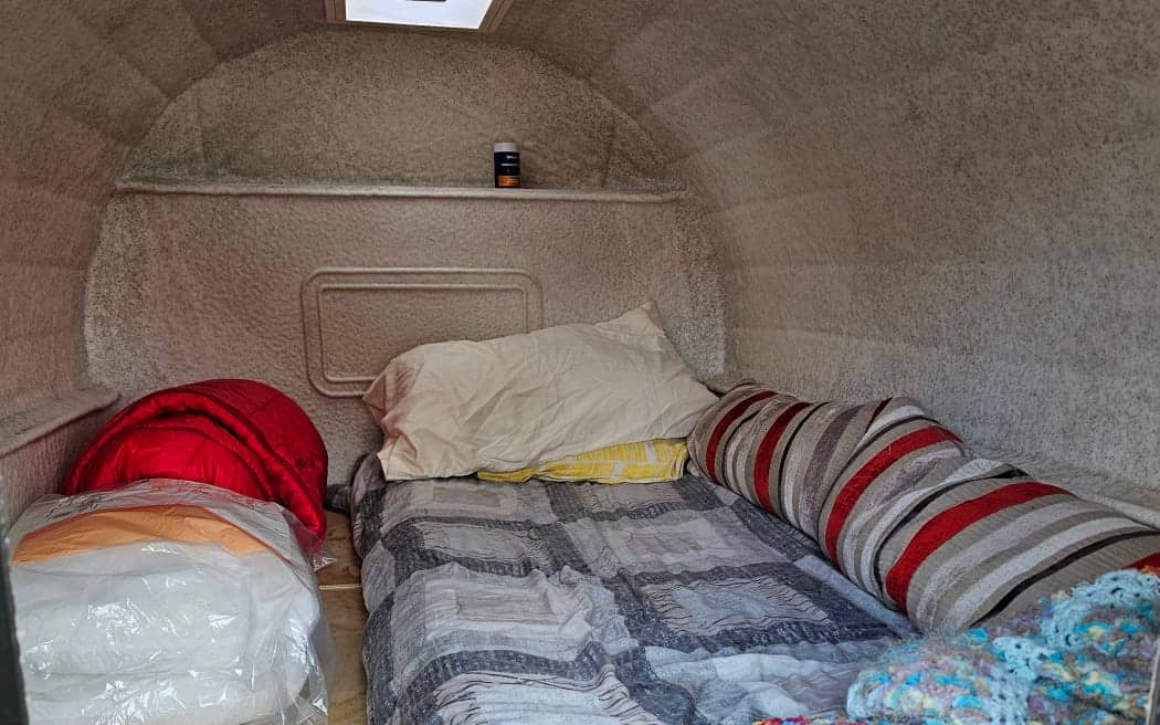A compact, insulated sleeping pod designed to provide shelter and warmth for homeless individuals. The interior features soft, beige textured walls and ceiling for insulation, with a neatly made bed covered by a gray and white patterned quilt, accompanied by a large pillow and a smaller yellow cushion. A striped cylindrical pillow rests against one wall, while a wooden bench on the side holds personal items including a red sleeping bag. A small skylight in the ceiling allows natural light to enter, creating a cozy and secure resting space aimed at offering comfort and protection in Rotorua’s challenging environment.