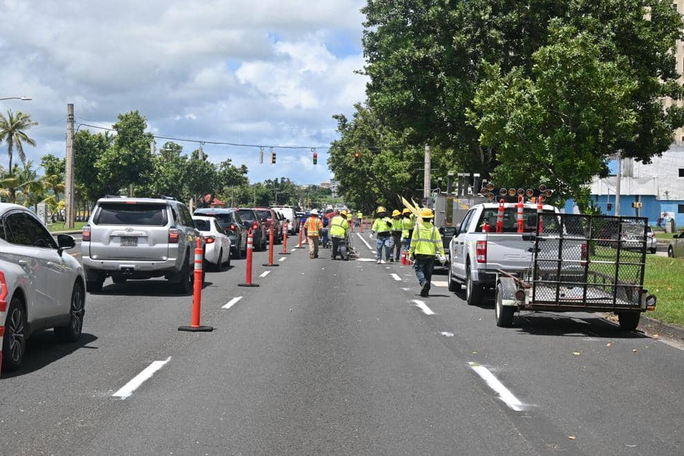 Road construction scene showing traffic cones, construction workers in high-visibility vests, and vehicles navigating around active roadwork on a paved street lined with tropical vegetation and palm trees in Guam.