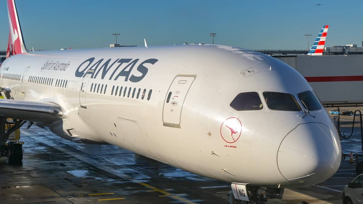 The nose section of a Qantas commercial airliner parked at an airport gate, showing the cockpit windows and part of the fuselage, with another Qantas aircraft tail visible in the background under a clear sky.