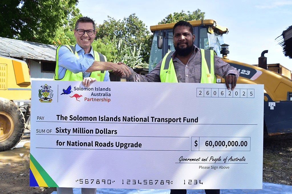  Two men in high-visibility safety vests shake hands while holding a large ceremonial check in front of yellow construction equipment. The check is made out to The Solomon Islands National Transport Fund for Sixty Million Dollars for National Roads Upgrade in the amount of $60,000,000.00, signed by the Government and People of Australia. The check features the Solomon Islands-Australia Partnership logo with both countries' emblems. The scene appears to be at a construction or road work site with trees in the background.