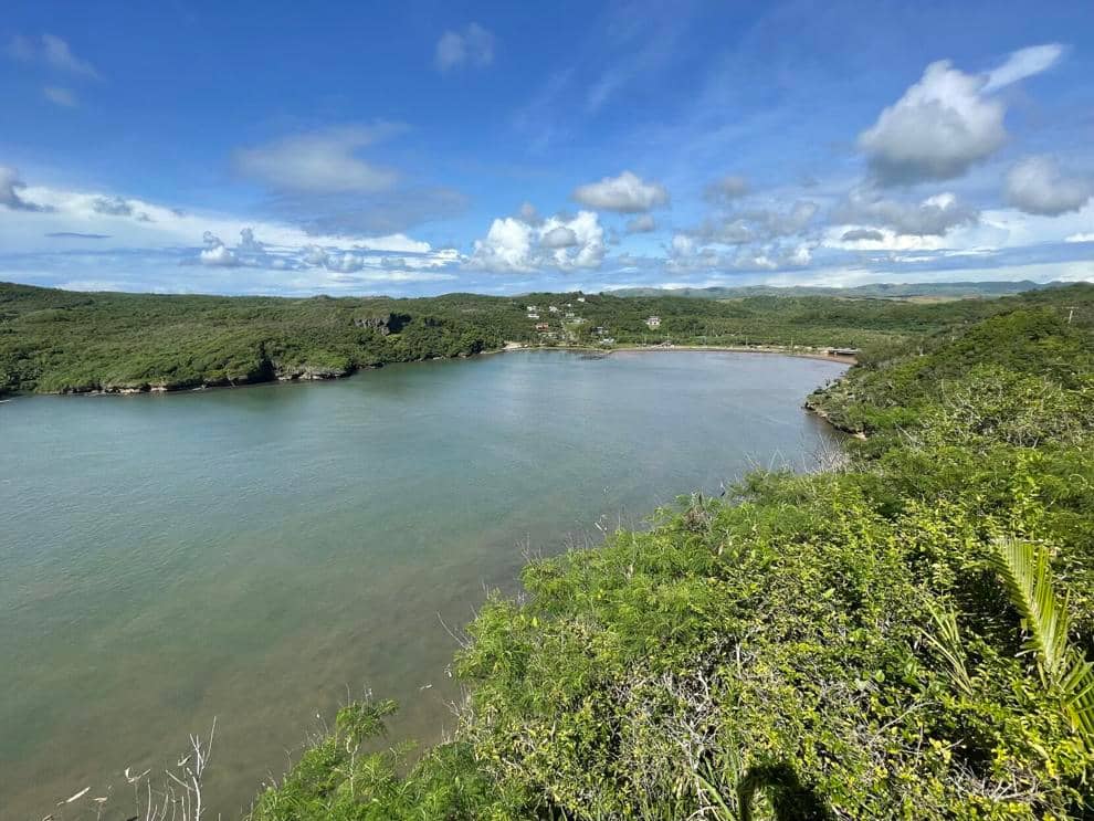 A panoramic view of a coastal inlet surrounded by lush green hills under a bright blue sky with scattered clouds. The calm, shallow water curves gently into the land, with a small village and road visible in the distance along the shoreline.