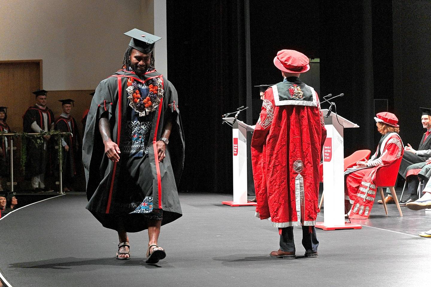 A person in academic regalia, Nikola Matawalu, featuring a black gown with red and blue accents and a lei around their neck, walks across a stage. He is wearing a graduation cap and sandals. Facing away from the viewer on the right side of the stage is another person in a red academic gown and hat, standing behind a white podium. Other individuals in academic attire are seated in the background.