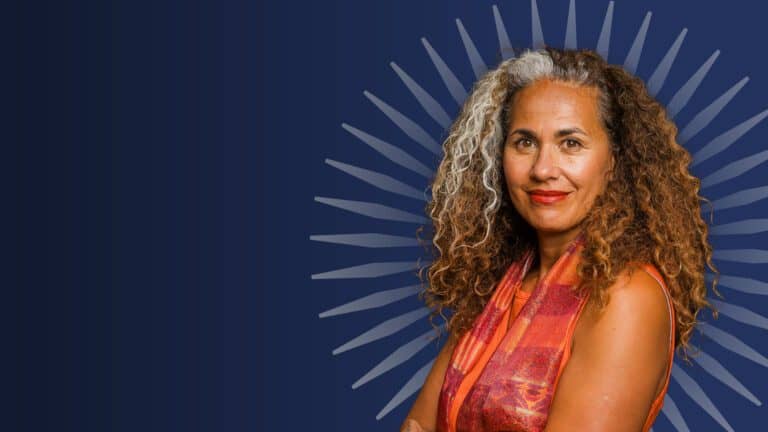 A portrait of Professor Selina Tusitala Marsh in front of a dark blue background. She is wearing a red and orange vest, and has her hair is let out in long light brown curls with grey edges in the front.