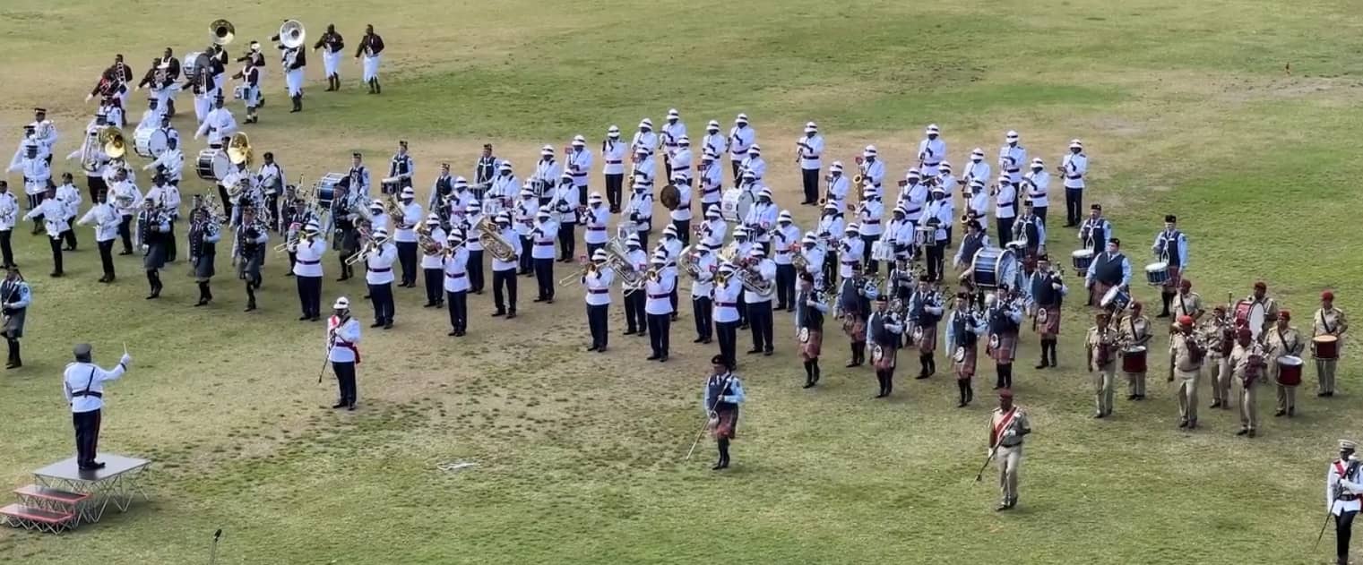 A large military band formation performing on a grassy field, with multiple groups in coordinated uniforms including white, light blue, maroon, and khaki. The musicians are playing brass instruments, drums, and bagpipes, while a conductor stands on a small platform at the front left directing the ensemble.” The room contains open cubby shelves, paperwork, and a window with curtains. The scene suggests a meeting or workshop related to a UNICEF-supported school meals initiative.