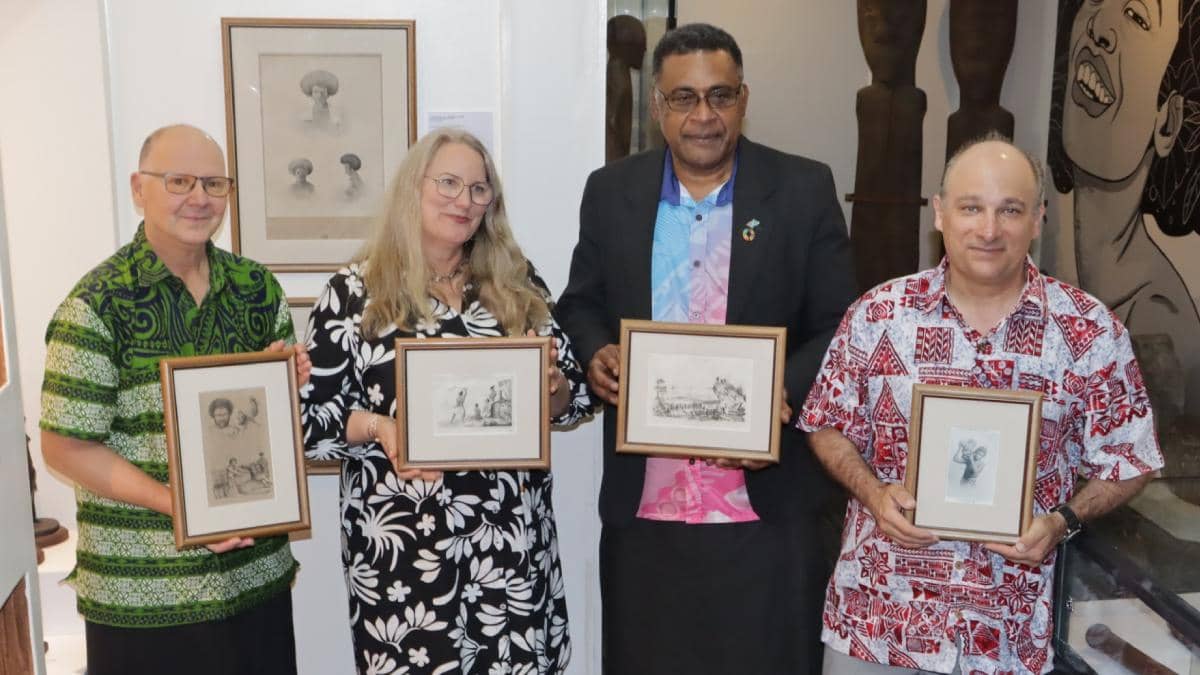 Four people standing in a museum or gallery setting, each holding framed historical photographs or artworks. From left to right: a man in a green patterned shirt, a woman with long blonde hair in a black floral dress, a man in a dark suit jacket, and a man in a red and white patterned shirt. They are positioned in front of white walls displaying various framed artworks and what appears to be traditional Pacific Island artifacts including carved wooden clubs. The setting suggests this is a formal presentation or donation ceremony at a cultural institution.
