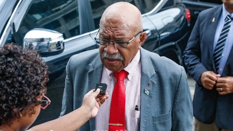 An older man wearing a grey suit, red tie, and striped shirt is being interviewed on the street. He has a bald head, glasses, and a white mustache, and is speaking into a small microphone held by a reporter. A black vehicle and another man in a suit are visible in the background.