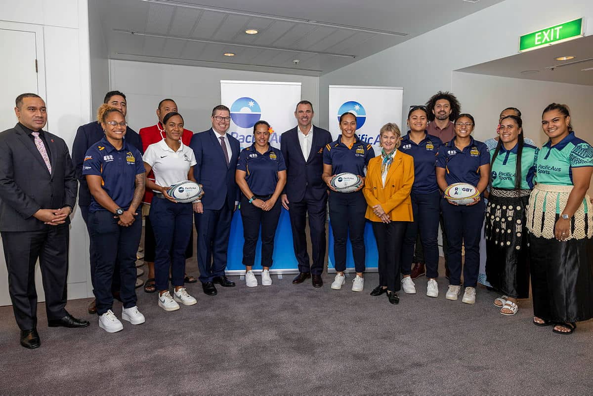 A group photo of approximately 15 people in business attire and rugby uniforms standing in a conference room, with several holding rugby balls, Pacific Aus banners visible in the background, and participants wearing various team jerseys including what app