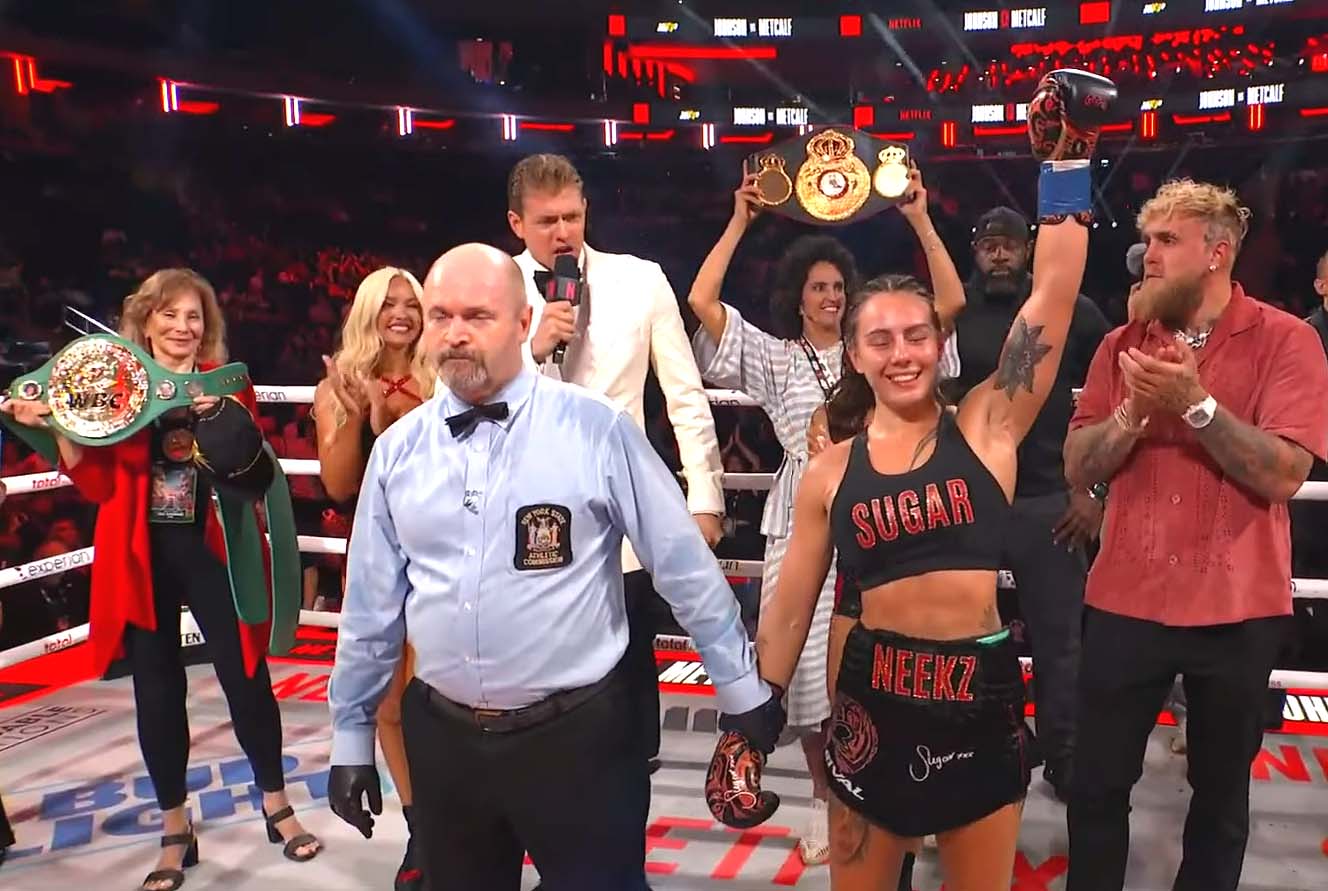 Boxing champion Cherneka Johnson celebrates her victory in the ring at Madison Square Garden, wearing black boxing shorts with 'SUGAR' and 'NEEKZ' printed on them. She stands with her arms raised in triumph, surrounded by officials, supporters, and what appears to be Jake Paul (in a red shirt) on the right. A referee in a light blue shirt is prominently featured in the foreground, and championship belts are visible being held up in the background. The scene is set against the iconic red and black Madison Square Garden backdrop with Netflix branding visible, capturing the moment after her historic TKO victory to become the first Māori undisputed boxing world champion.