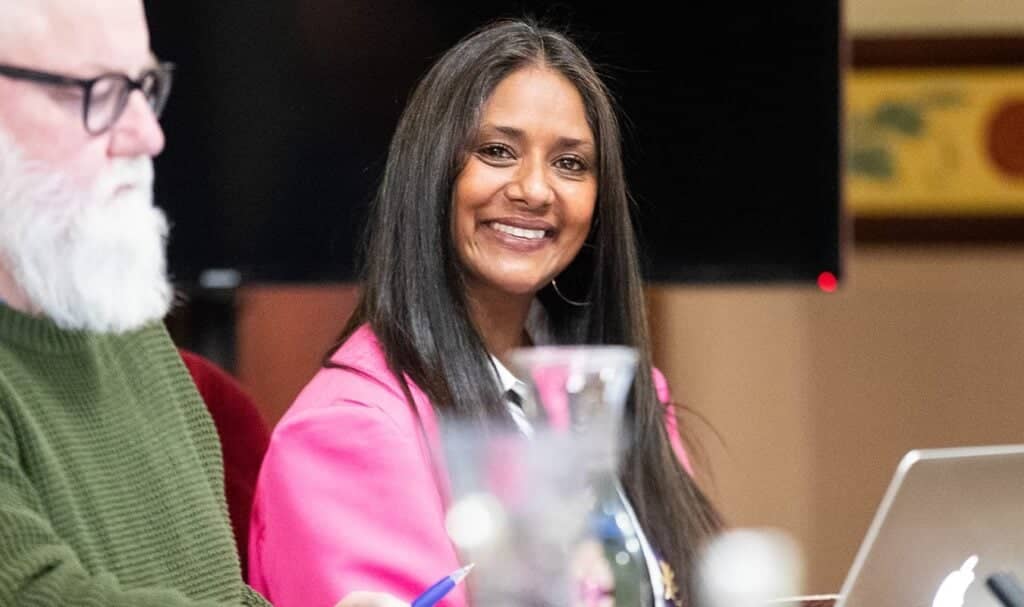 A person with long dark hair wearing a bright pink jacket, Dr Zelinda Sherlock, sits at a table with a laptop and glass water jug in front of them. Another individual in a green sweater is seated nearby. The setting appears to be a meeting or conference room with a dark monitor and artwork on the wall in the background.