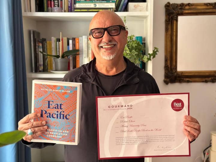 A smiling bald man with glasses and a goatee, Robert Oliver, holds up the Eat Pacific cookbook in his left hand and the official Gourmand World Cookbook Awards certificate in his right hand. He is standing in front of a white bookshelf filled with books, with a framed mirror visible on the wall behind him.