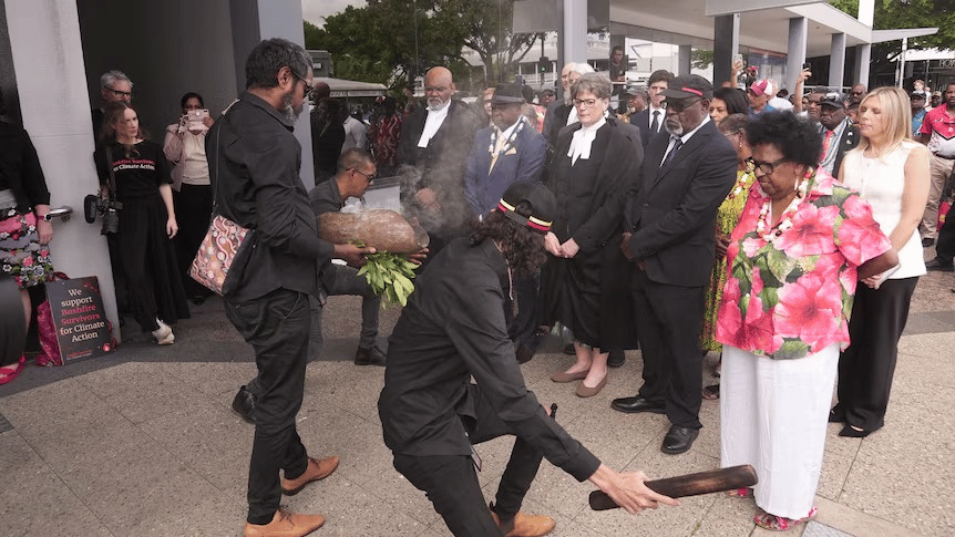 A ceremonial gathering takes place outdoors, where two men in black clothing perform a traditional Aboriginal smoking ceremony with eucalyptus leaves and a smoking wooden bowl. One man crouches with a clapping stick while another holds the smoking vessel. A group of people—including elders, officials in formal attire, and onlookers—stand respectfully in a semicircle observing the ritual. A woman in a vibrant pink floral blouse and white pants stands near the front. In the background, others are taking photos and watching the event. A sign on the left reads: We support businesses for climate action.