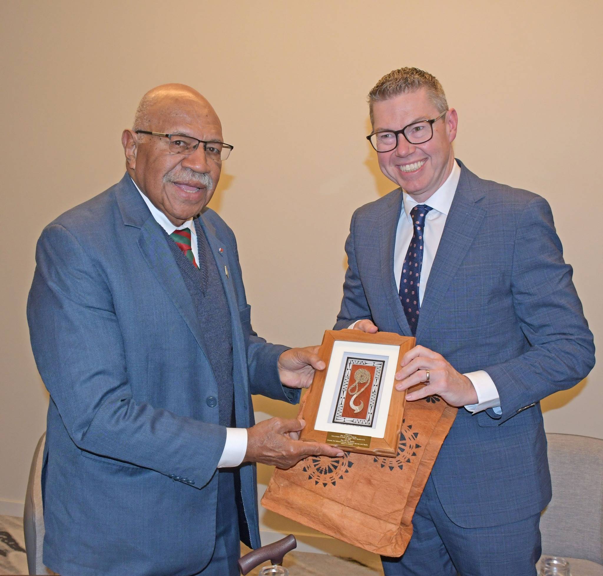 Two men in dark blue business suits shake hands while holding a wooden plaque or award between them. The man on the left is older with glasses, a mustache, and is bald. The man on the right is younger with glasses and light-colored hair. They are standing in front of a neutral beige background and both are smiling at the camera during what appears to be a formal ceremony or meeting.