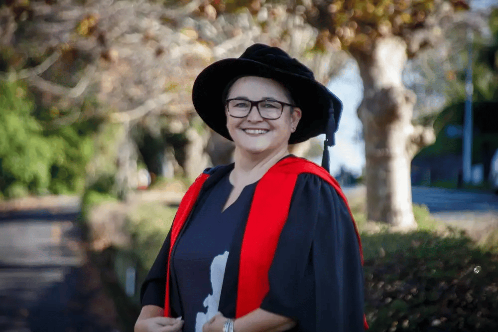 Dr Lara Tookey is wearing academic regalia, including a black gown with red trim and a traditional academic cap, stands outdoors on a tree-lined path during autumn.