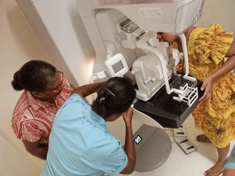 Two healthcare workers assist a woman in a yellow patterned dress who is positioned at a mammography machine for a breast screening procedure in a medical clinic.
