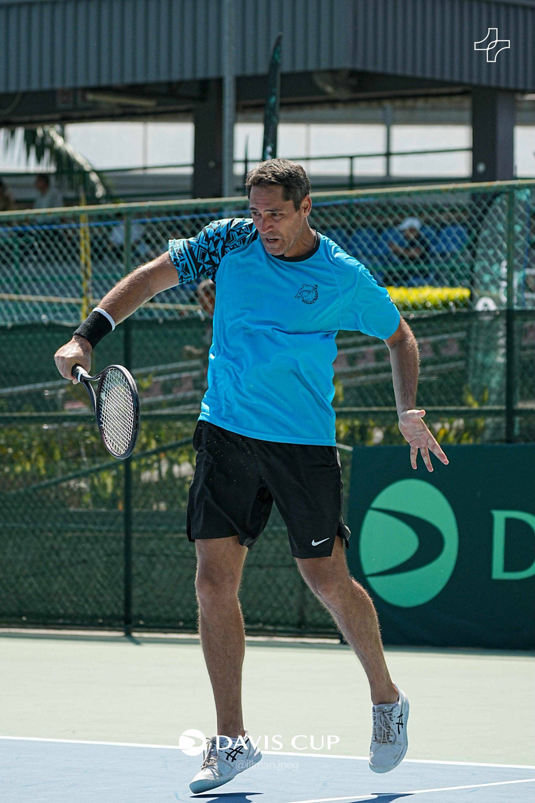 A male tennis player in a blue shirt and black shorts, holding a tennis racket, is captured mid-action on a green outdoor court with a 
