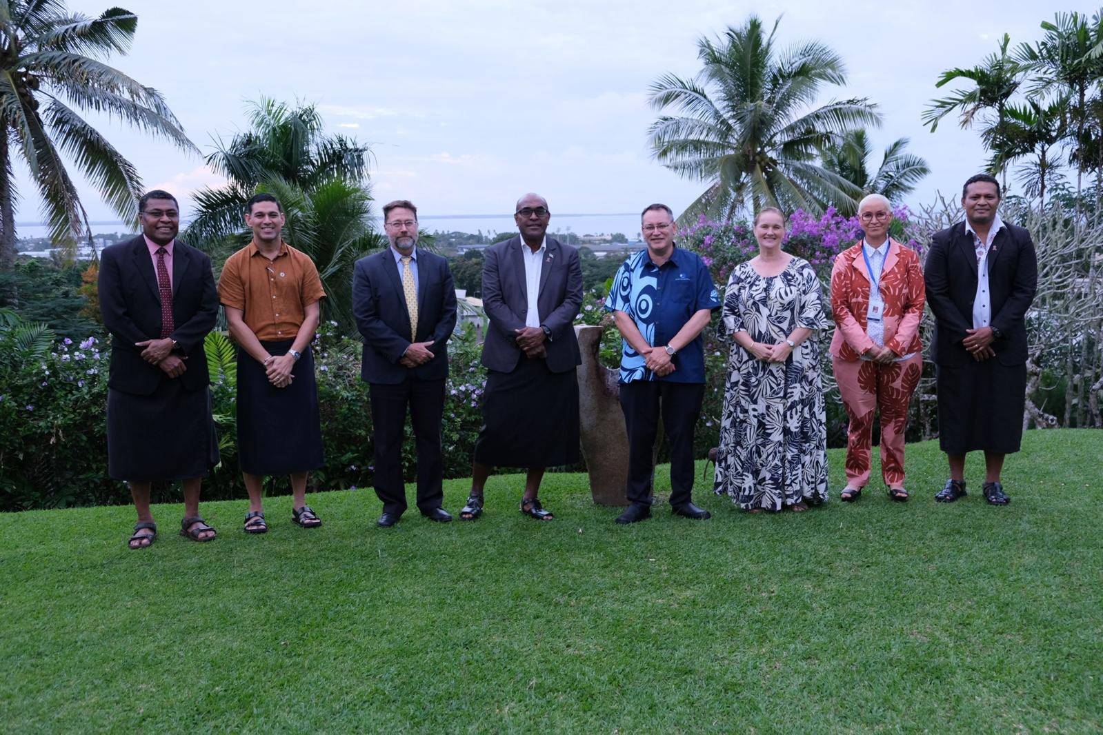 A group of eight individuals stands in a line on a green lawn with tropical plants and palm trees in the background. The people are dressed in a mix of formal attire and traditional clothing, with some wearing patterned shirts, skirts, or suits. The setting appears to be outdoors in a lush, tropical environment, possibly overlooking a distant town or coastline.