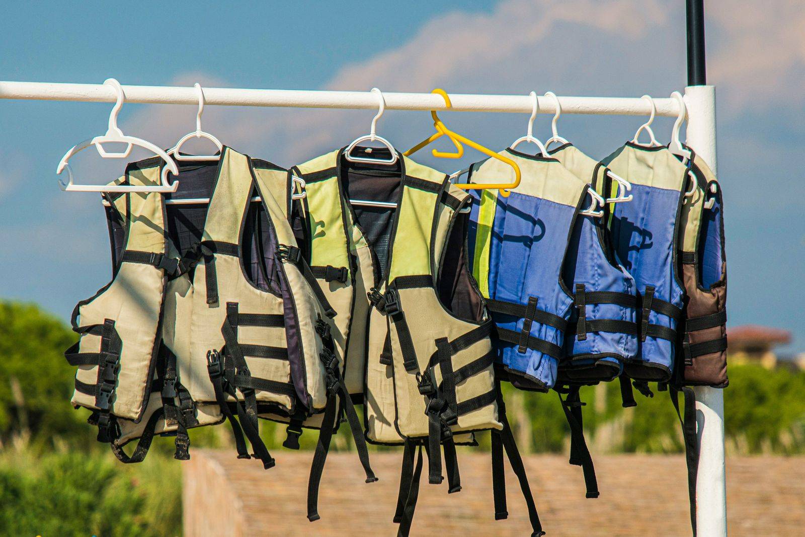 Multiple personal flotation devices and lifejackets hanging on white hangers from a white rod to dry outdoors. The lifejackets are in various colors including beige, yellow, blue, and gray, all with black safety straps and buckles. They're photographed against a blue sky with white clouds, with green vegetation visible in the blurred background.