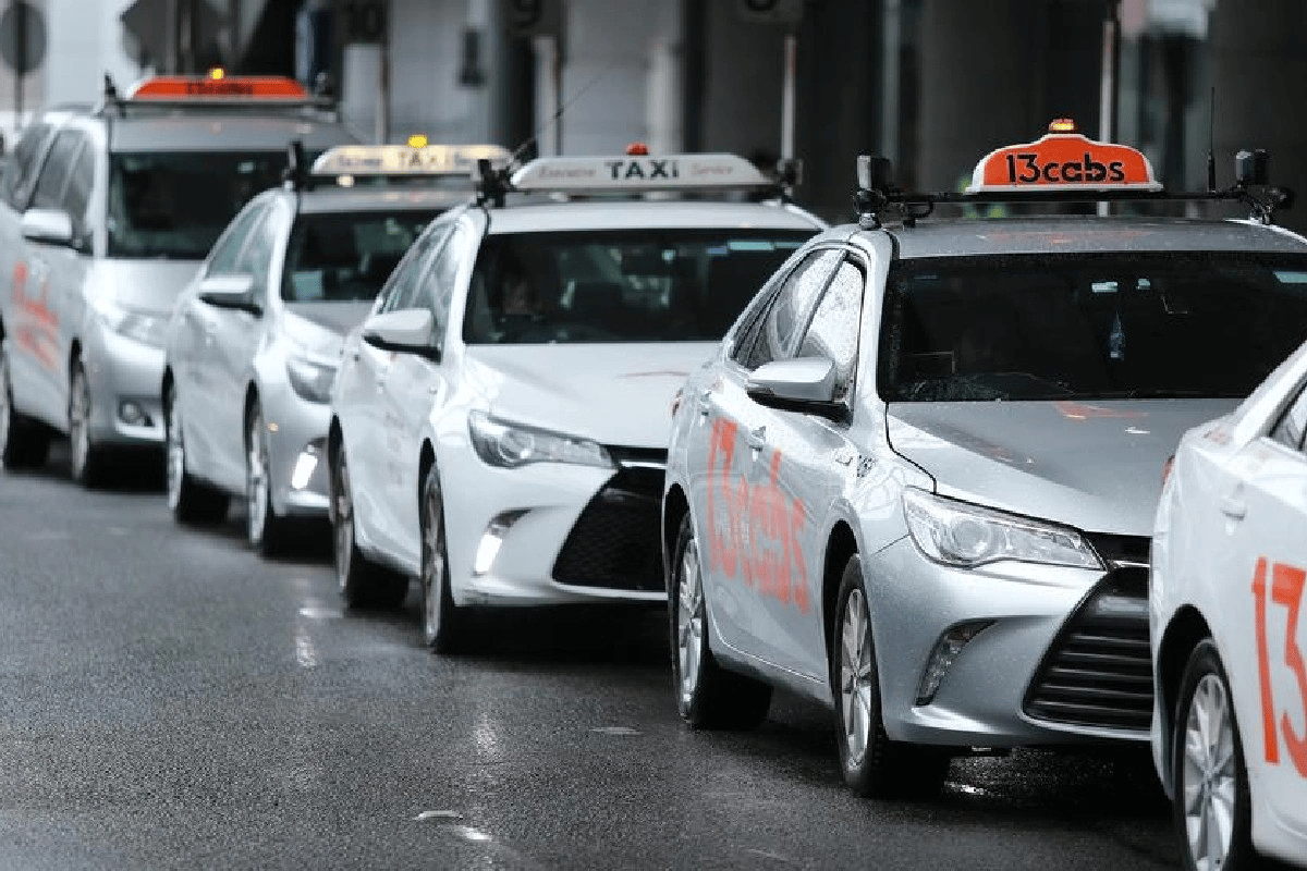 A line of white and silver taxis, likely Toyota Camrys, is parked on a wet street. Each taxi has a yellow TAXI sign on its roof, with some also displaying 13cabs or a similar numerical identification. The cars are facing slightly to the right, and the wet pavement suggests it might have recently rained or is currently raining.