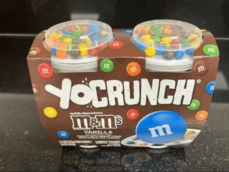 A product shot of a YoCrunch M&M's Vanilla Lowfat Yogurt two-pack. The brown cardboard sleeve features the YoCrunch logo in white, with colorful M&M's scattered across it. Two clear plastic cups of yogurt are visible at the top, each with a clear lid containing a separate compartment filled with milk chocolate M&M's for mixing into the yogurt. An oversized blue M&M candy is prominently displayed on the front, with 'm' printed in white on it. The background is a dark, possibly countertop, surface.