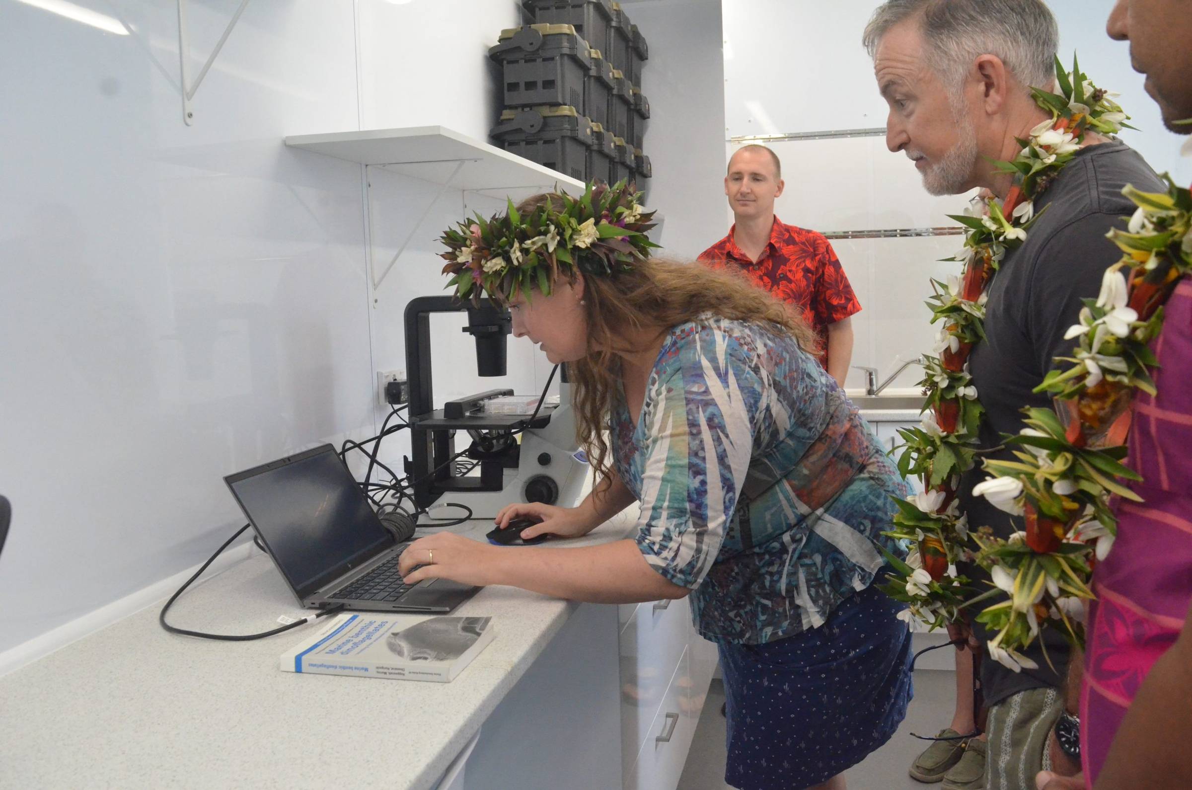 A woman wearing a flower crown leans over a laptop connected to a microscope in a laboratory setting. She is demonstrating or analyzing something on the screen, while several people, also adorned with floral garlands, observe attentively.