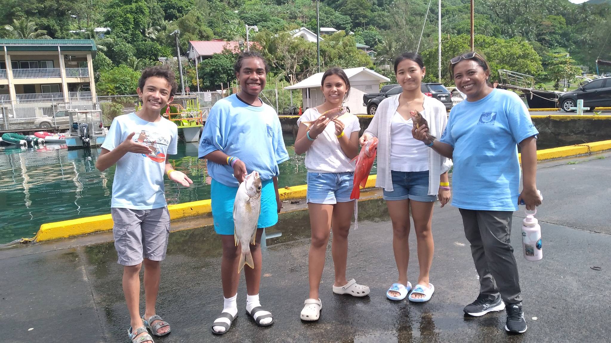 A group of five people, including an adult woman and four children/teenagers, stand on a wet dock holding fish they've caught. They are all smiling, with boats and green hills visible in the background.