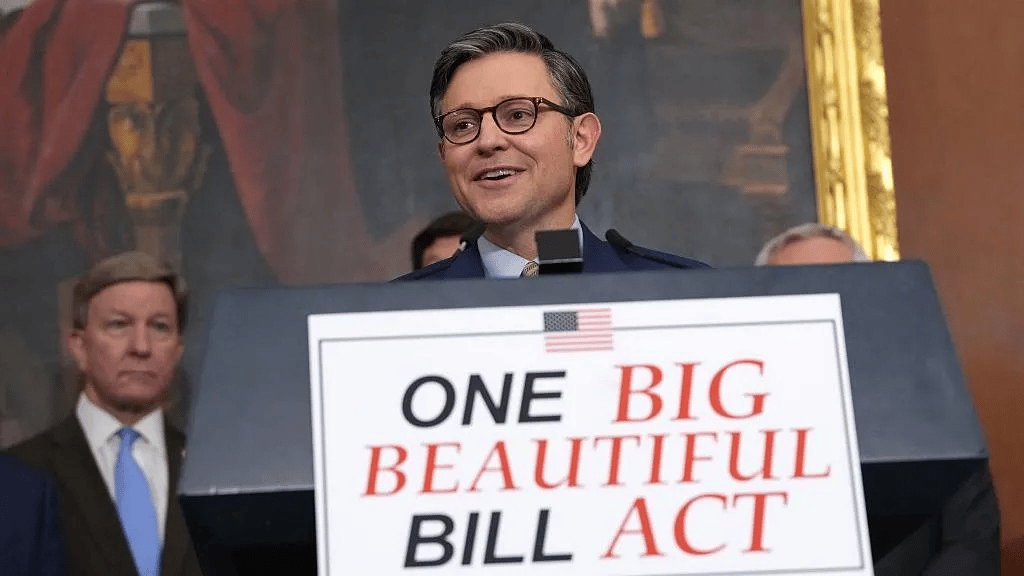 A man, House Speaker Mike Johnson, in glasses and a dark suit speaking at a podium with a sign reading 'ONE BIG BEAUTIFUL BILL ACT' with an American flag symbol, standing in what appears to be an ornate government building with other officials visible in the background