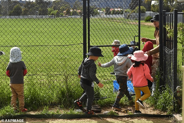 Children dressed in warm clothes and hats being led by an adult woman through a dark grey swing gate into a large open grassy field.
