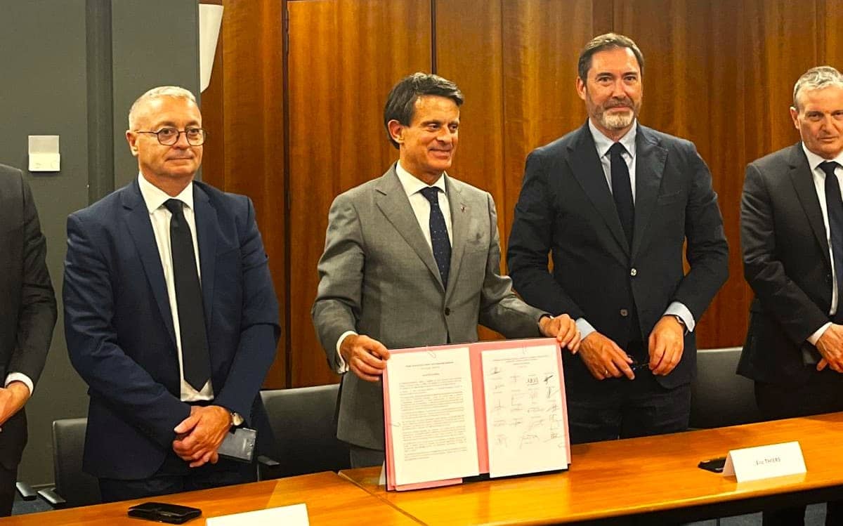 Four men in dark business suits stand behind a wooden conference table in a formal meeting room. A signed document with a red ribbon is prominently displayed on the table in front of them. The men appear to be posing for a ceremonial photo following what appears to be a formal agreement signing. The setting suggests an official government or diplomatic meeting.