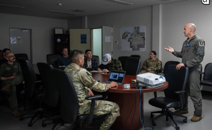 Military briefing room scene showing a uniformed officer standing and presenting to a seated group of military personnel around a conference table. The standing officer appears to be giving a presentation or briefing, with his hand extended in a gesturing motion. Approximately 6-8 military personnel in various camouflage uniforms are seated around a dark wooden conference table, with laptops and documents visible on the table surface. The room has gray walls, fluorescent lighting, and appears to be in a military facility or command center. Maps or charts are visible on the walls in the background.