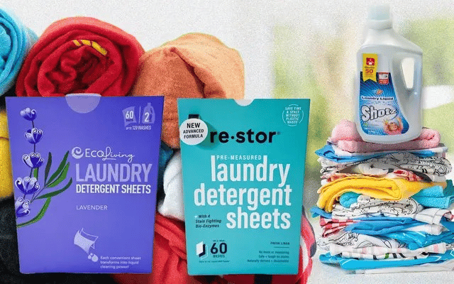 A comparison display of laundry products: two boxes of eco-friendly laundry detergent sheets (one lavender-scented by EcoLiving and the other fresh linen-scented by Re:stor) are placed against a background of colorful folded towels. To the right, a bottle of traditional liquid detergent labeled Shooz sits on top of a neatly folded stack of clean laundry.