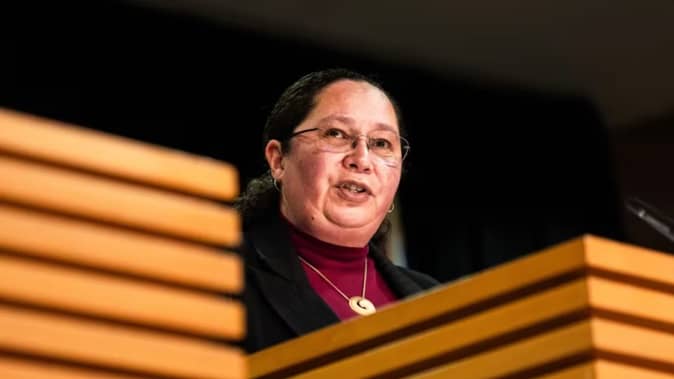 Associate Justice Minister Nicole McKee speaking at a wooden podium during a press conference, wearing glasses and a purple top with a necklace.