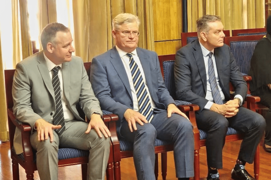Three men in business suits sit side by side on wooden chairs with blue cushions in a formal indoor setting, featuring gold curtains and wood-paneled walls.