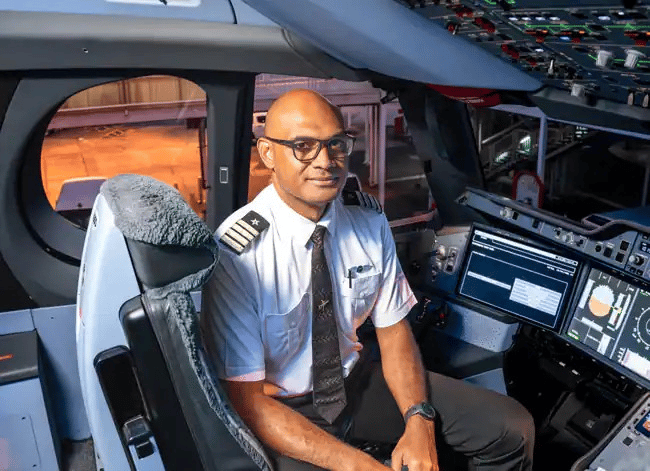 A smiling bald man with glasses, wearing a white pilot's shirt with epaulets, a dark tie, and a watch, sits in the cockpit of an airplane. He is looking directly at the viewer. The cockpit is visible around him, with various screens and controls.