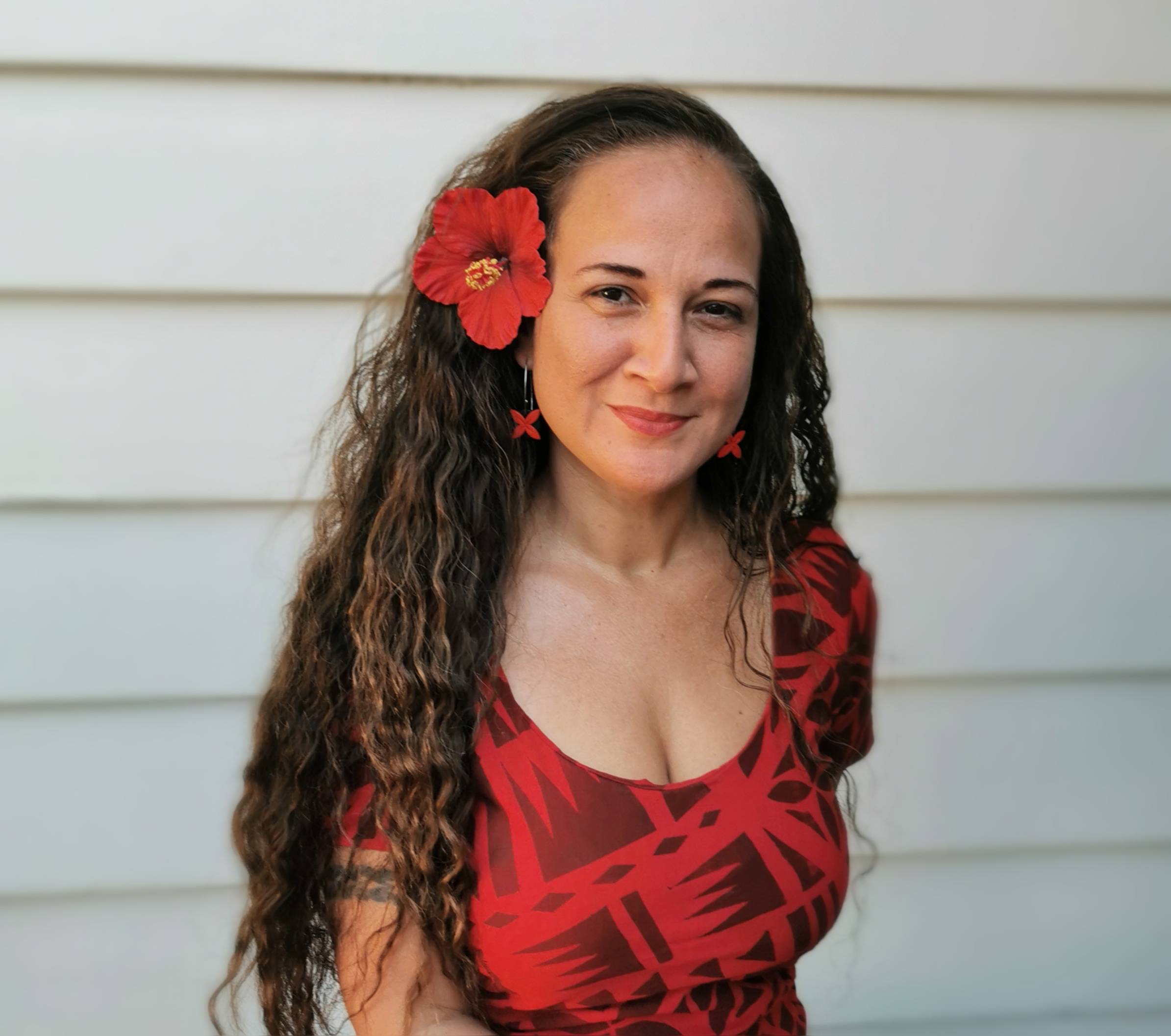 A woman, Dr. Lagipoiva Cherelle Jackson, with long, curly dark hair smiles warmly at the camera while wearing a vibrant red hibiscus flower behind her left ear. She's dressed in a red top with a traditional Pacific Islander geometric pattern and matching 