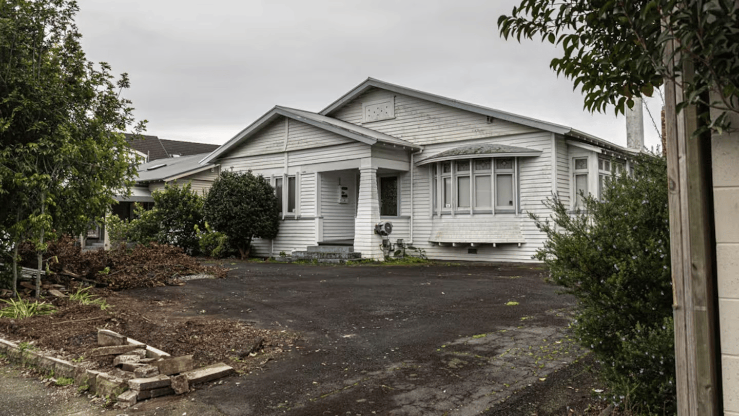 A weathered white wooden house with a gabled roof sits under a grey overcast sky. The house features a covered front porch, bay windows, and visible air conditioning units. The front yard is partially cleared, with scattered bricks, trimmed bushes, and patches of bare soil. The driveway and surrounding vegetation suggest the property is undergoing maintenance or renovation.