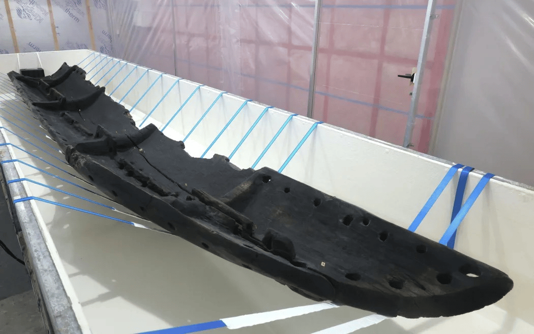A well-preserved ancient canoe, dark and weathered, is displayed in a white support cradle with blue straps in a controlled environment. The canoe’s hull is lined with large holes and internal supports, indicating its historic craftsmanship and maritime u