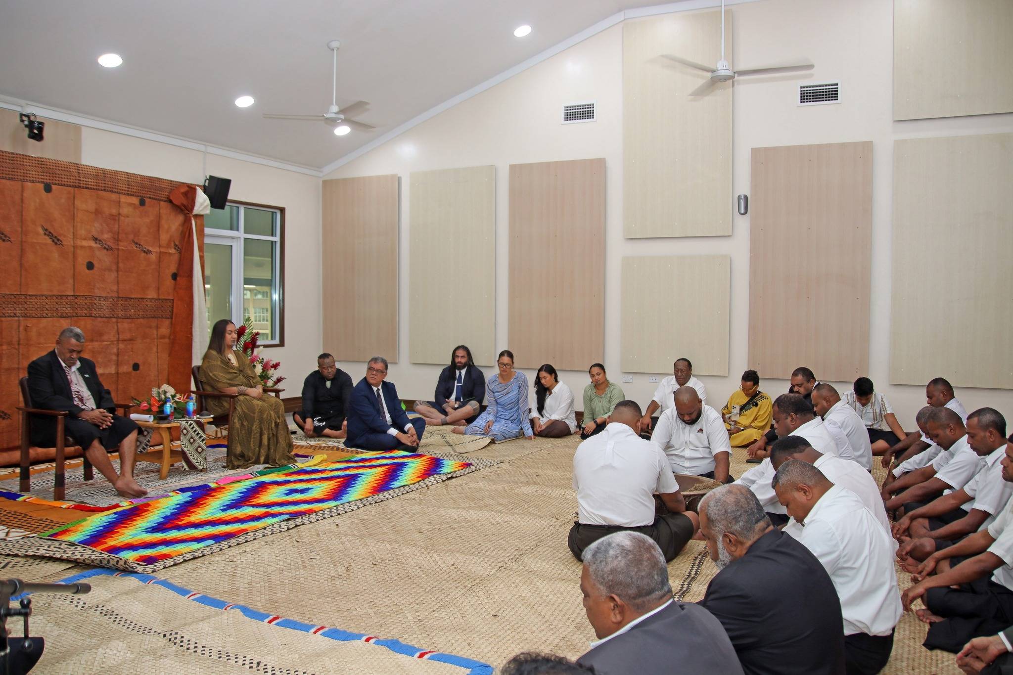 A woman in a patterned brown dress, Her Majesty Te Arikinui Kuini, the M?ori Queen, walks barefoot across a colorful woven mat in a room decorated with tapa cloth, tropical flowers, and traditional Fijian furnishings, during a formal cultural ceremony.