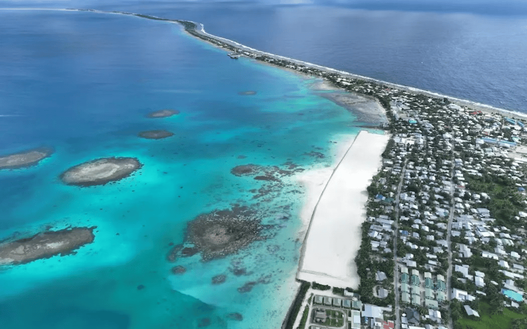 Aerial view of a low-lying Pacific island atoll, Tuvalu, with turquoise waters, coral reefs, a white sandy airstrip, and dense residential settlement on a narrow strip of land between lagoon and ocean.