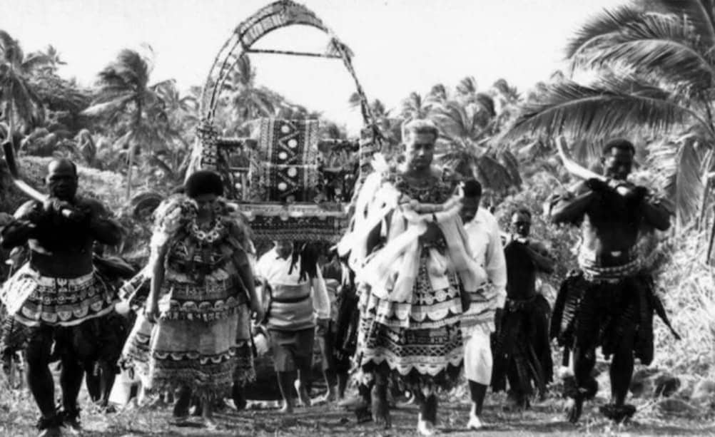 A black-and-white photograph showing a traditional Fijian procession. Several people are dressed in elaborate, patterned ceremonial attire, some carrying a large, ornately decorated structure on their shoulders. Coconut trees and tropical vegetation are visible in the background, suggesting a festive or significant cultural event in an island setting.