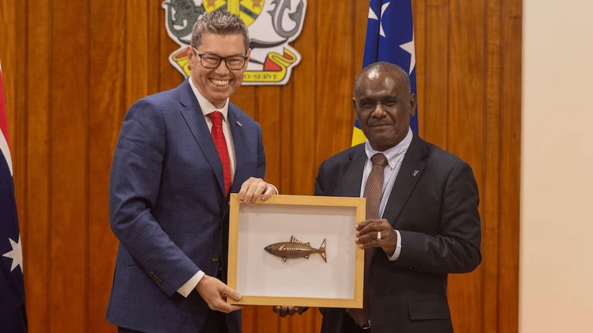 Two men, Pat Conroy and Jeremiah Manele, in business suits stand together in what appears to be a government office, holding a framed gift featuring a golden fish ornament. The man on the left wears glasses, a blue suit with red tie, and is smiling broadly. The man on the right wears a dark suit with patterned tie. Behind them are visible government emblems and flags, including what appears to be a Solomon Islands flag with its distinctive blue field and yellow stars. The setting suggests an official diplomatic meeting or ceremony.