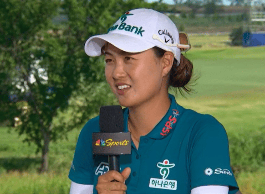 Minjee Lee being interviewed on the golf course, holding a microphone with the lush green fairway visible in the background.