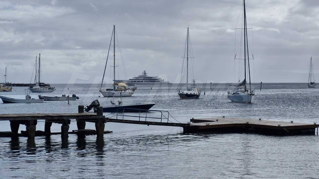 A harbor scene showing multiple sailboats and yachts anchored in calm waters under a cloudy sky, with a wooden dock and small motorboat in the foreground and a large luxury yacht visible in the background.