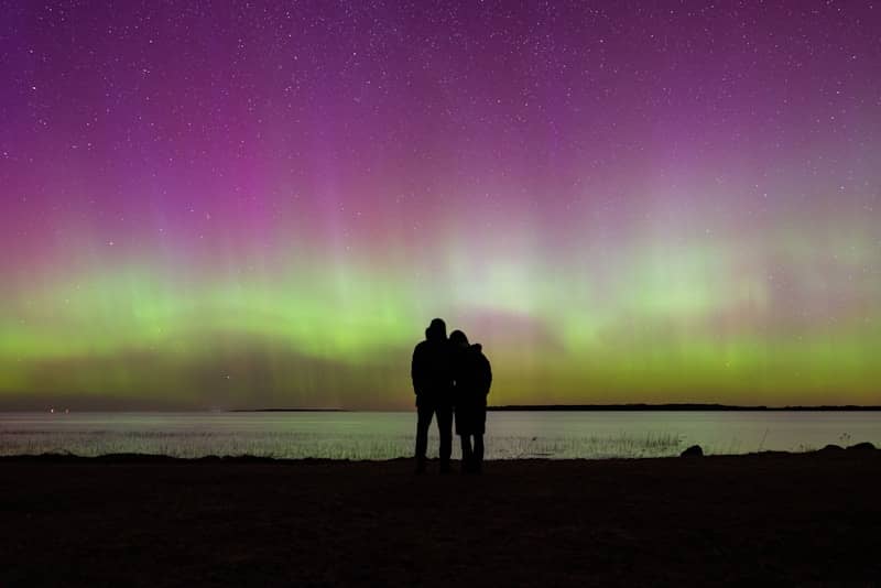 Couple watches the aurora borealis over water.
