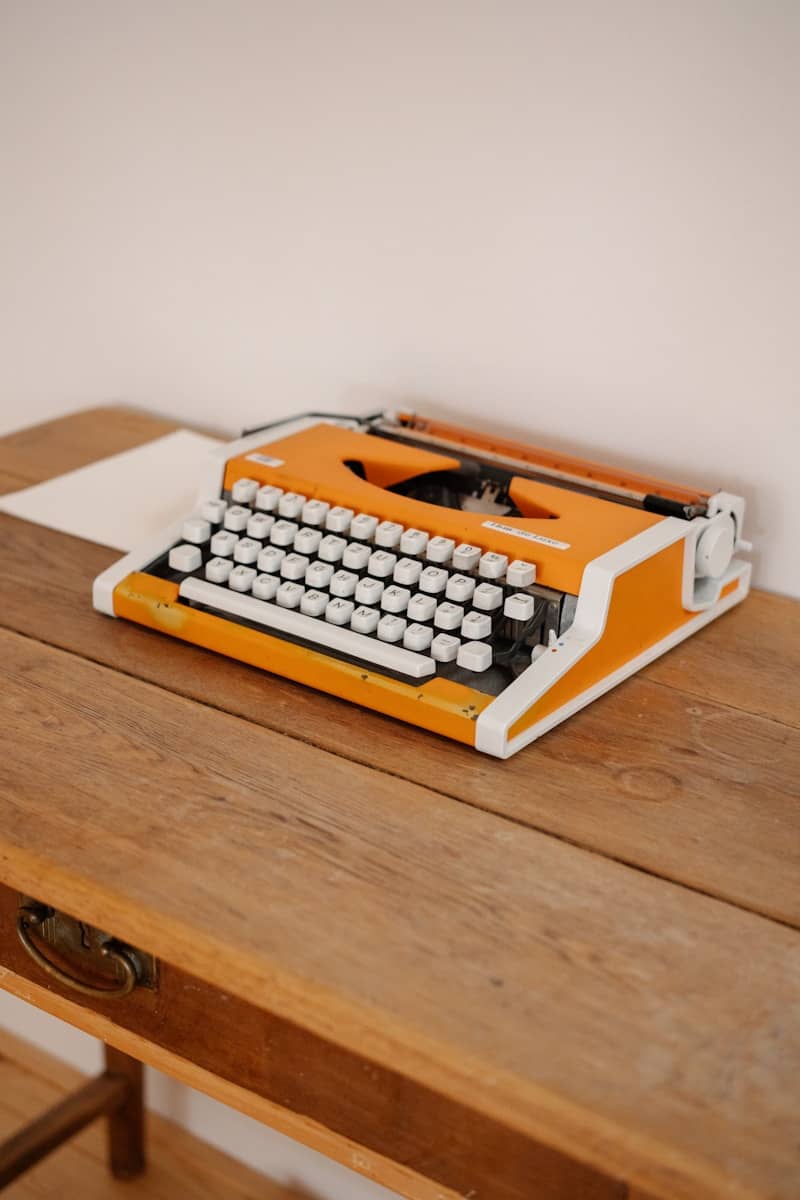 An orange typewriter sits on a wooden desk.