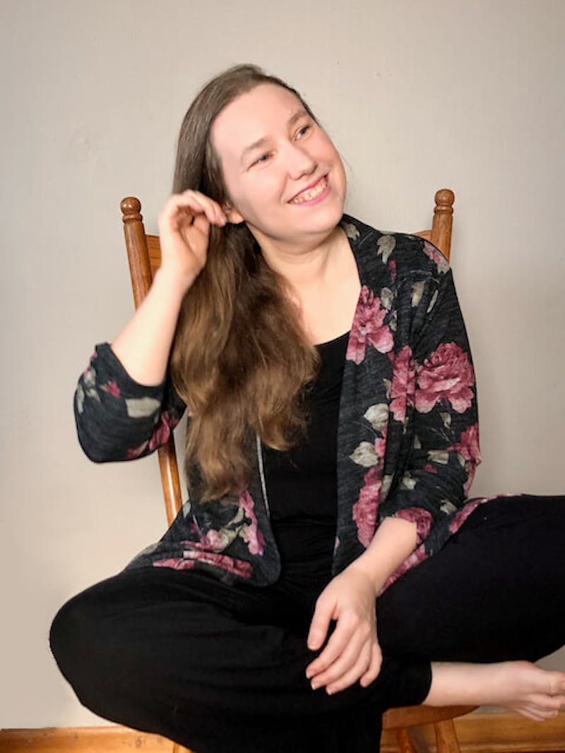 Woman smiling and wearing a shirt with roses and black pants. She has pale skin and long brown hair pulled over her right shoulder, sitting in a chair with legs crossed and tugging at her right ear.