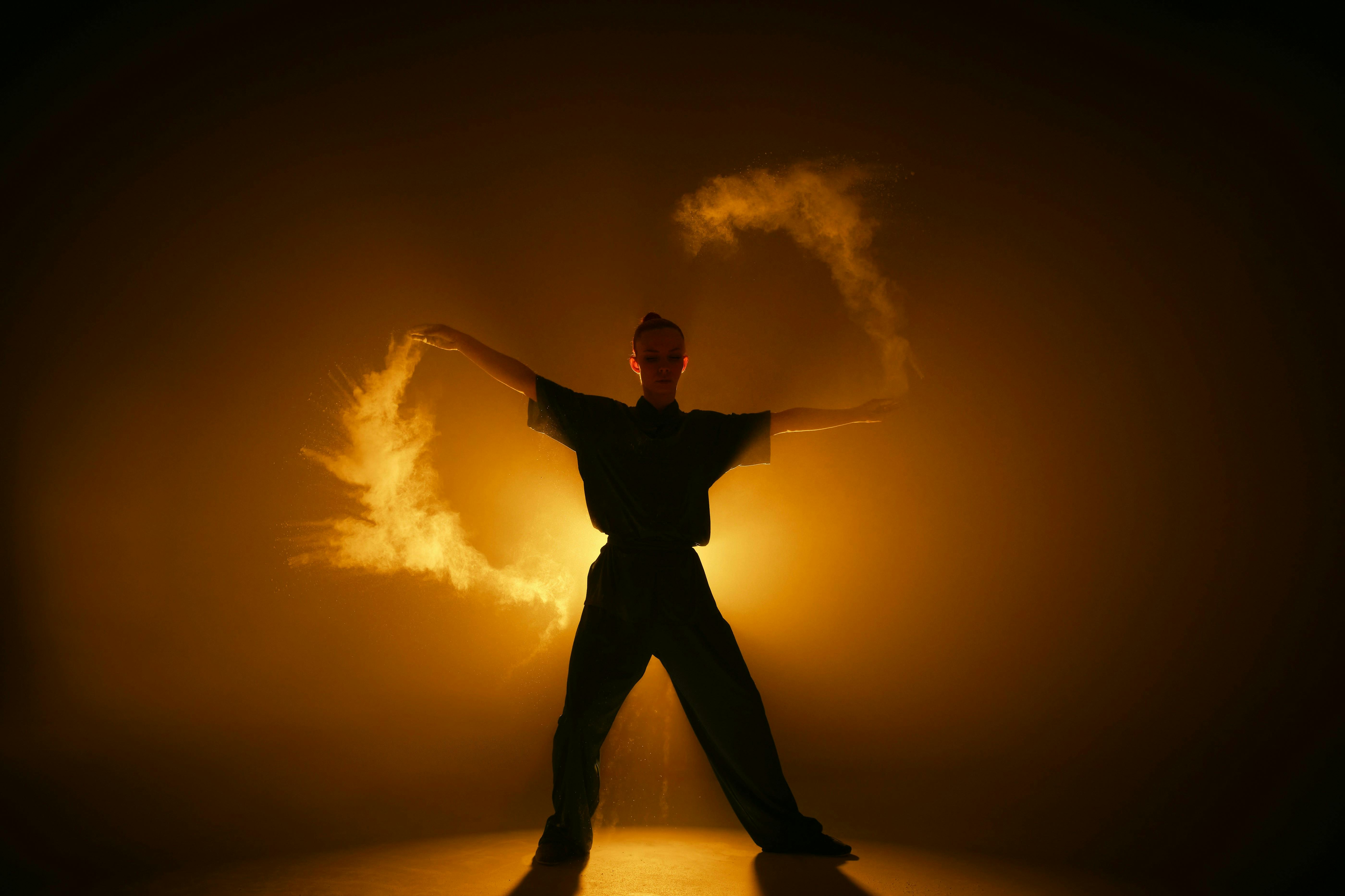 A black silhouette figure standing with arms and legs spread, flinging yellow powder in a ring around themself, backlit in yellow light.