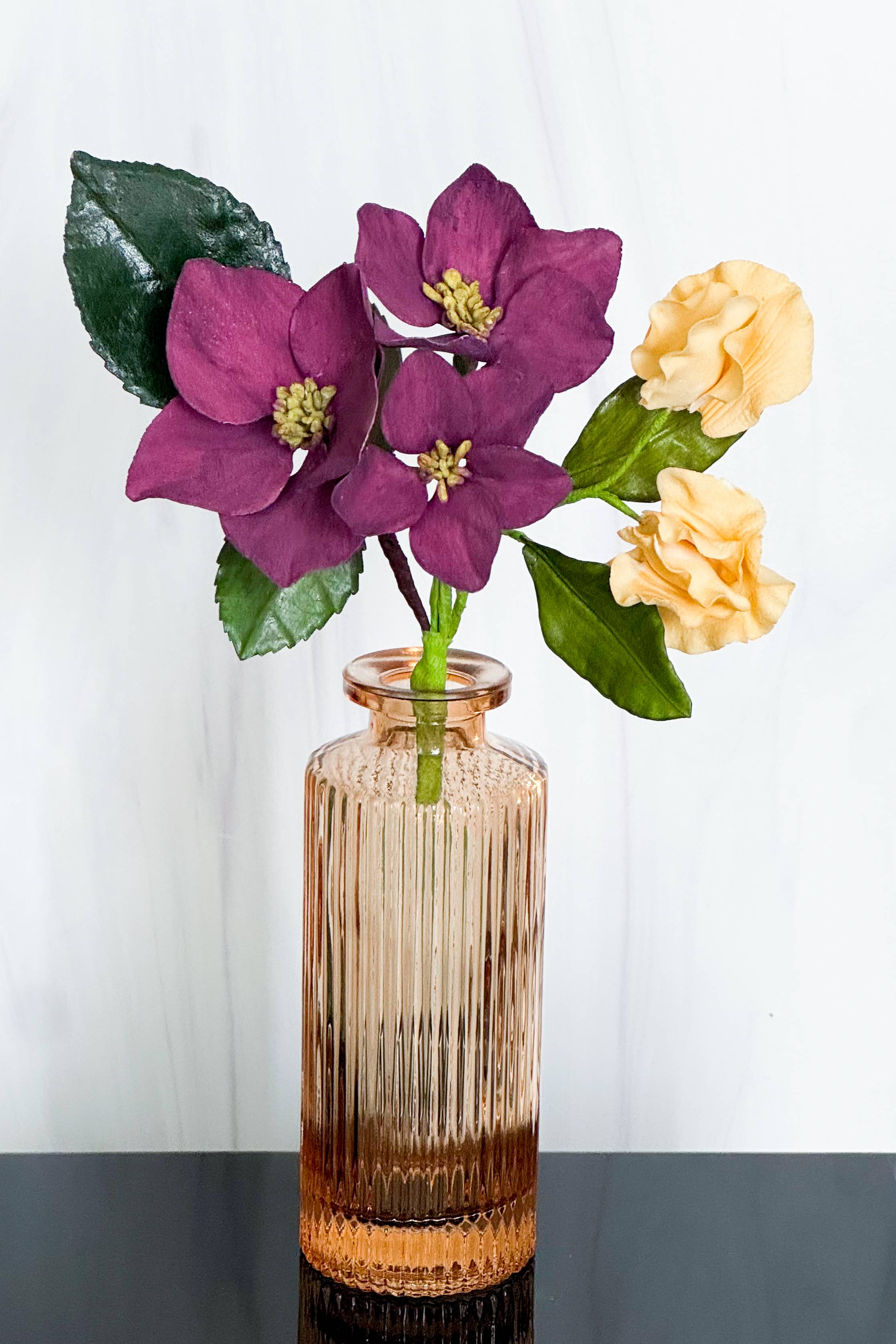a peach colored vase filled with sugar flowers like dark purple hellebores, peachy orange sweet peas, and dark green foliage against a plain background