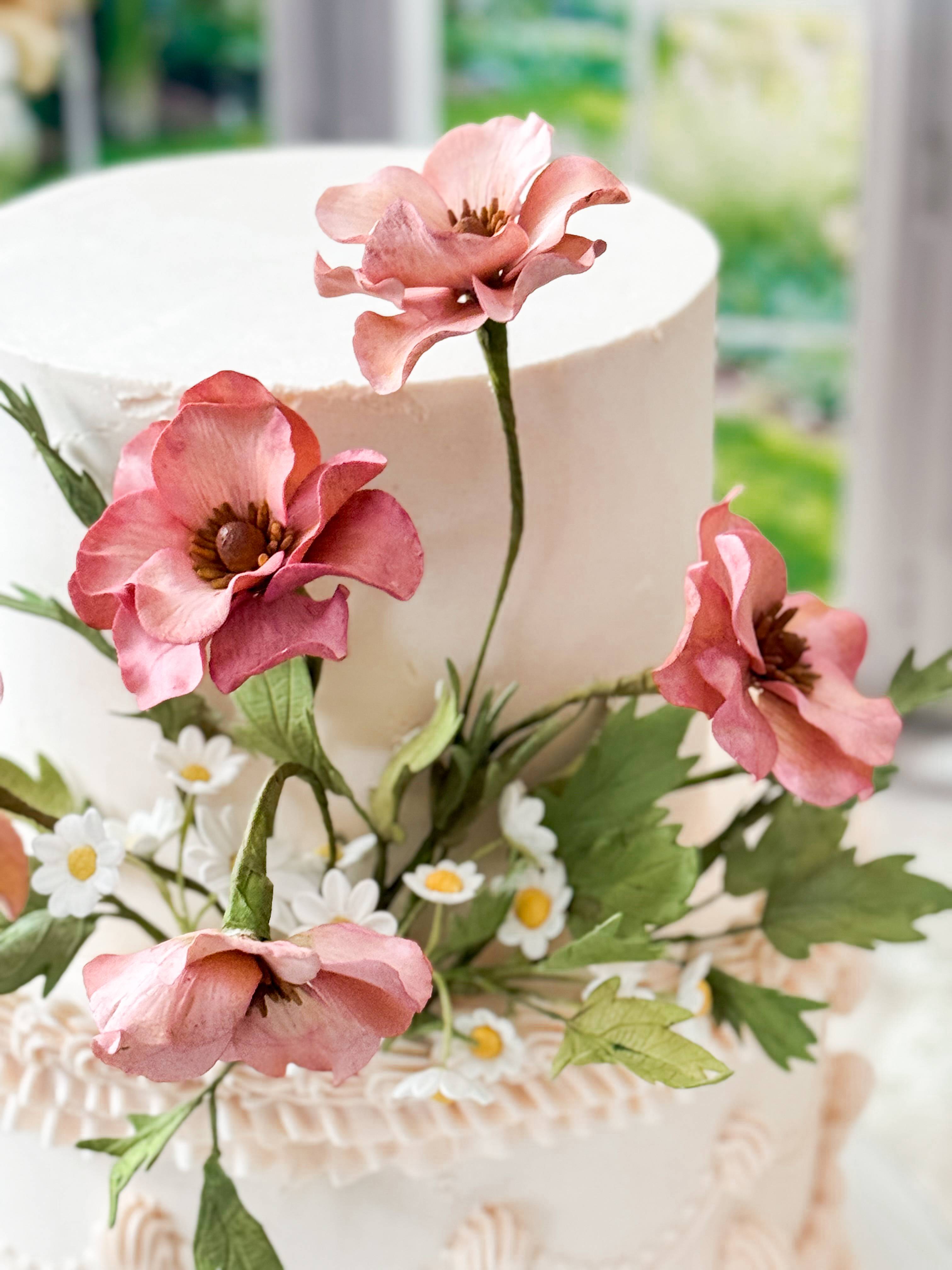 close up of pink sugar flowers with light green leaves and white and yellow daisies on a two tier pale pink buttercream cake